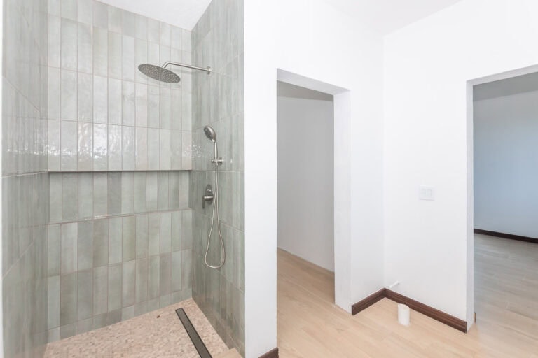 A modern bathroom with a glass-enclosed shower featuring a rain showerhead and handheld shower, set against greenish tiles typical of Honduras and Roatan homes.