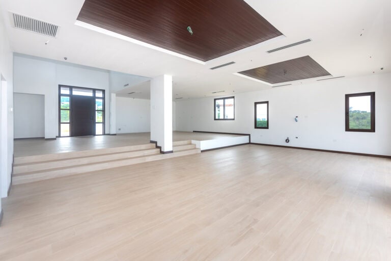 Spacious empty interior room in a modern building on Roatan, Bay Islands, Honduras, featuring wooden floors, large windows, and a high ceiling with wood panel accents.
