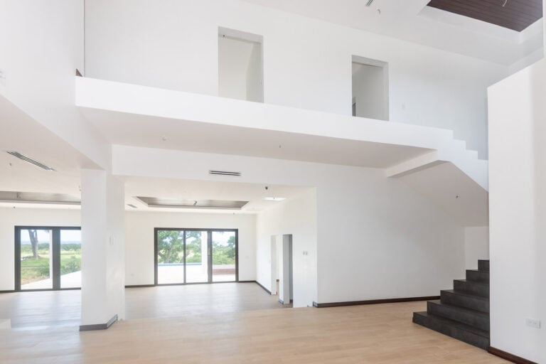 The image shows a spacious, modern interior of a house in Roatan, Honduras, with white walls, large windows, and a staircase leading to an upper level, overlooking scenic views of the surrounding landscape.