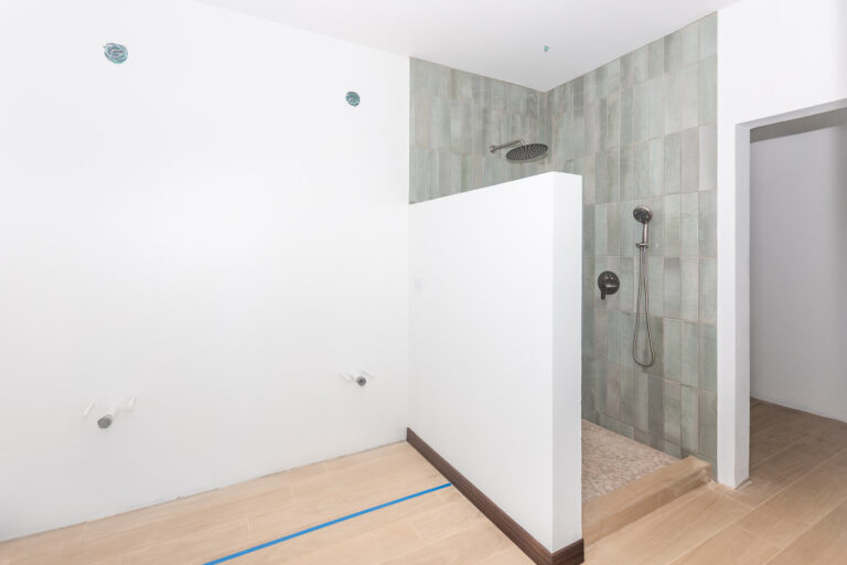 A modern indoor shower area on Roatan, Honduras with a partial wall separating the open shower space, featuring a ceiling-mounted rain showerhead, a handheld showerhead, and light green tiles on the wall.
