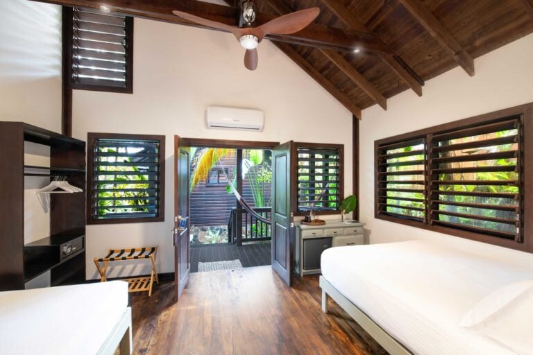 A bright, airy bedroom in Roatan, Honduras, features wooden ceiling beams, multiple louvered windows with tropical greenery outside, and simple white furniture including beds and a small dresser, with a hammock visible through the open door.