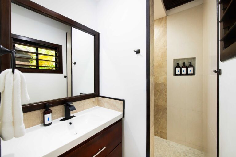 A modern bathroom in Roatan, Honduras features a wooden-framed mirror above a white vanity with black fixtures, a towel hanging on a hook, and a separate shower area with beige tiled walls and three bottles of toiletries placed in a niche.