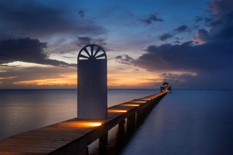 A white open door with a semi-circular window stands at the beginning of a wooden pier illuminated by small lights along its edge, extending into calm waters with a small gazebo at the end, set against a colorful sunset sky over Roatan in Honduras.