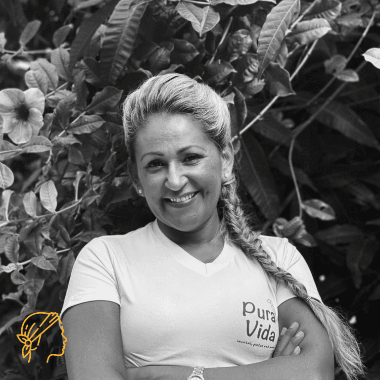 A smiling woman with a long braid stands outdoors in Roatan, Honduras, surrounded by lush foliage and large leaves.