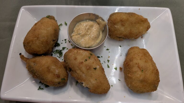 Four golden-brown breaded fish fillets served on a white rectangular plate with a small metal cup of tartar sauce in the center, garnished with chopped parsley, on a table in Roatan, Honduras.
