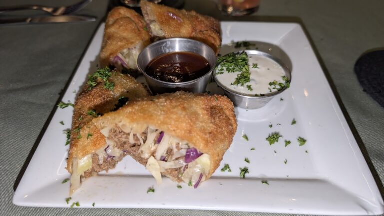 Close-up of a plate with crispy battered and fried spring rolls filled with vegetables and meat, accompanied by dark soy sauce and a creamy white dipping sauce garnished with chopped herbs, served on a white square plate in Roatan, Honduras.