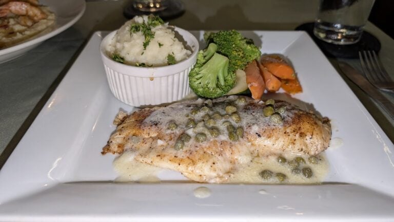 Grilled fish fillet served with creamy caper sauce, accompanied by mashed potatoes, steamed broccoli, and roasted carrots on a white square plate at a restaurant in Roatan, Honduras.