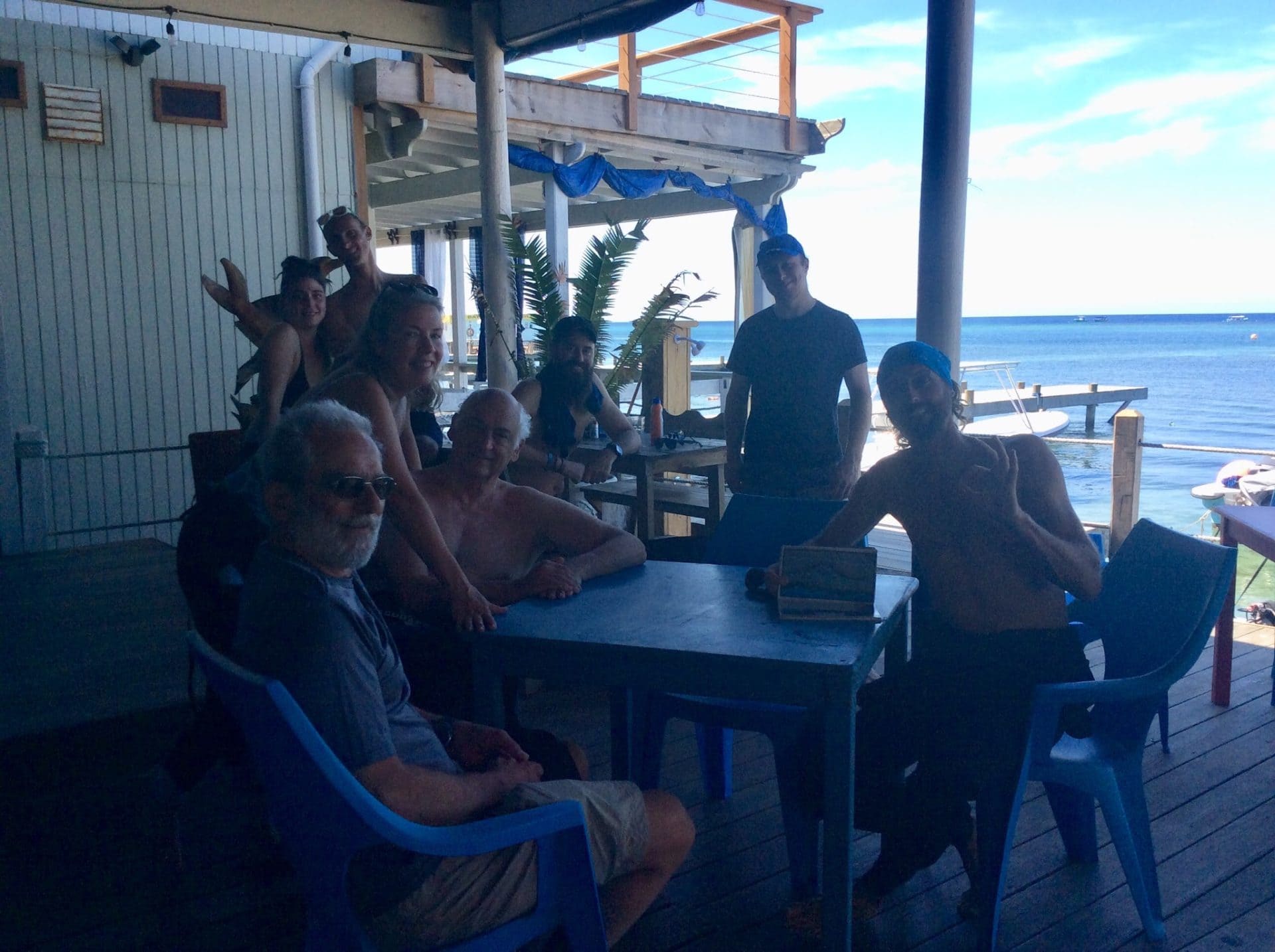A group of people relax and enjoy drinks at an outdoor restaurant or bar on the waterfront in Roatan, Honduras, with the ocean and boats visible in the background.