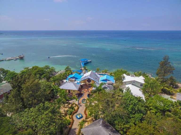 Aerial view of a resort with blue-roofed buildings, lush greenery, and a dock extending into the turquoise waters of Roatan in Honduras.