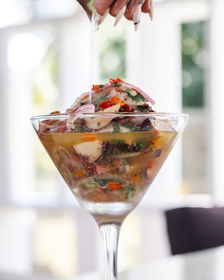 A person is pouring a colorful seafood ceviche into a glass martini glass on a table in Roatan, Honduras, with a blurred bright background.