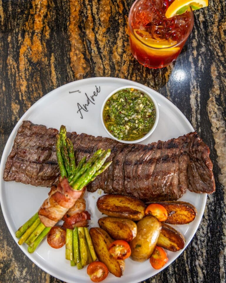 A plate of grilled steak, roasted potatoes with cherry tomatoes, bacon-wrapped asparagus, and a small bowl of chimichurri sauce is served on a white dish with the restaurant name "André" in Roatan, Honduras, with a glass of iced cold red drink garnished with lemon and lime on a dark, patterned table.