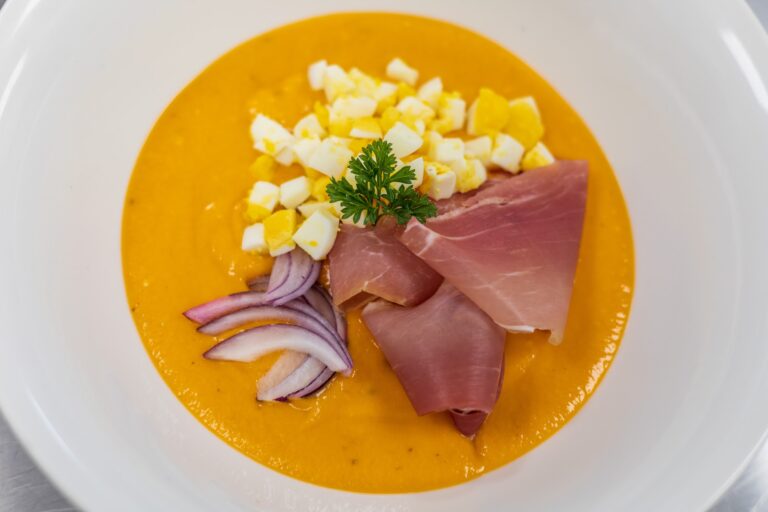 A bowl of vibrant orange pumpkin soup topped with chopped hard-boiled egg pieces, sliced red onion, a sprig of parsley, and slices of ham, served in a white dish on Roatan, Honduras.