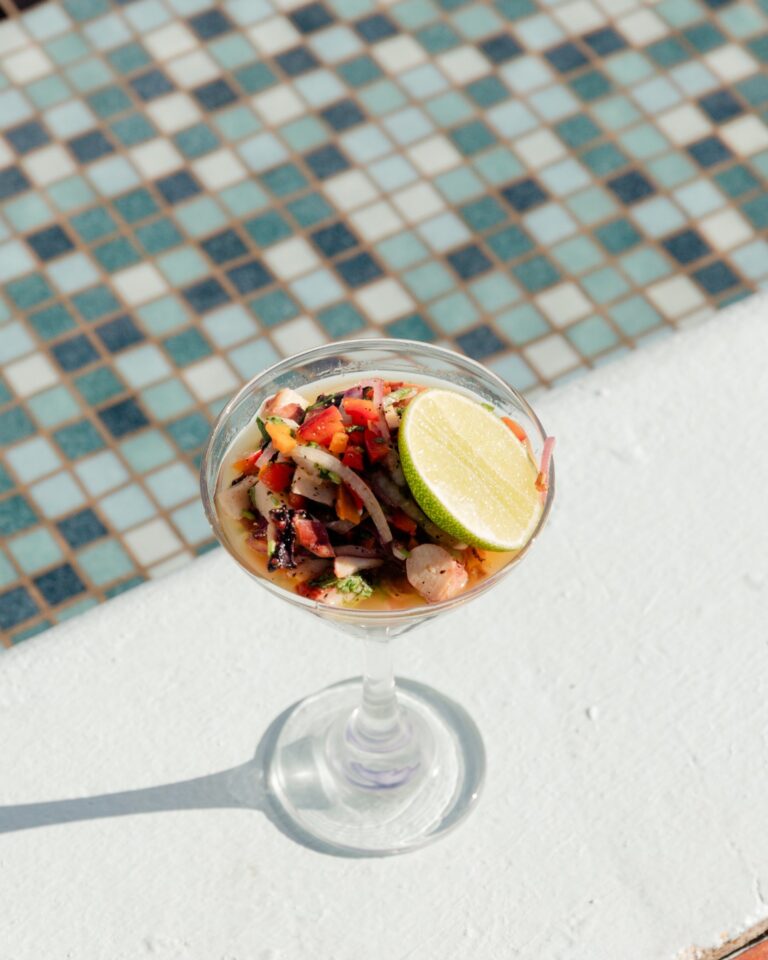 A bowl of ceviche with diced seafood, vegetables, and herbs, garnished with a lime wedge, is placed on a white surface next to a swimming pool with small, multicolored mosaic tiles in Roatan, Honduras.