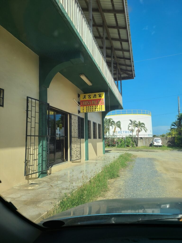A view from inside a vehicle showing a building with a sign for China Palace, a business in Roatan, Honduras, with palm trees and a Petro gas station visible in the background under a clear blue sky.