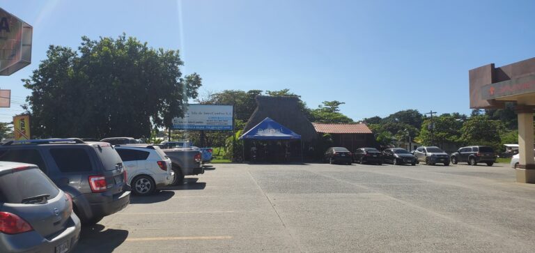 A busy parking lot in Roatan, Honduras, featuring a blue Pepsi tent, various parked cars, and signs for local businesses amid trees and clear blue sky.