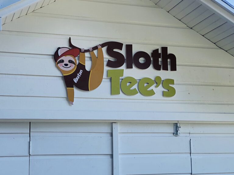 A colorful sign featuring a smiling sloth wearing a red cap and holding a hammock, with the words "Sloth Tees" on a white wooden wall in Roatan, Honduras.