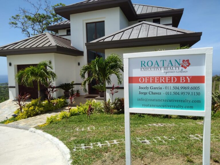 A modern two-story house with white exterior walls, dark metal roof, and large windows, situated in a lush landscaped garden with palm trees and colorful plants, is displayed alongside a sign for Roatan Executive Realty in Roatan, Honduras.