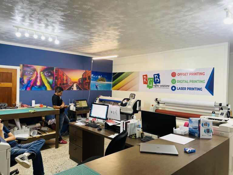 The image shows an indoor business office or printing shop in Roatan, Honduras, with various graphic design and printing equipment, colorful artwork on the walls, and employees working at their desks.