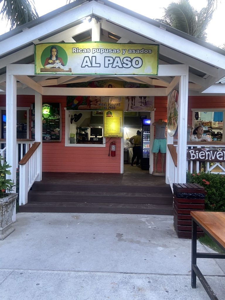 The image shows the entrance to a small food stand on Roatan, Honduras, with a sign advertising delicious roasted and grilled pig parts, and a person working inside.