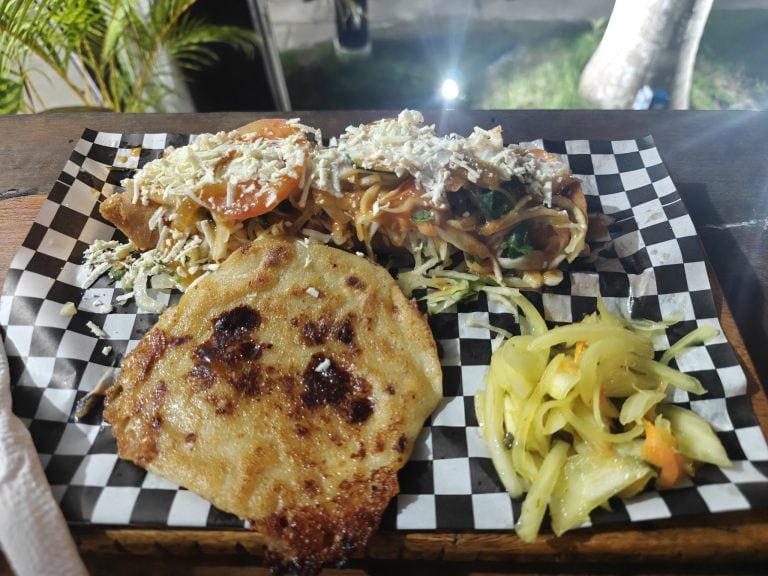 A plate of Mexican-style food, including a cheese and vegetable quesadilla, topped with shredded cheese, served with pickled cabbage, and other toppings, set on a checkered paper in a dining area in Roatan, Honduras.