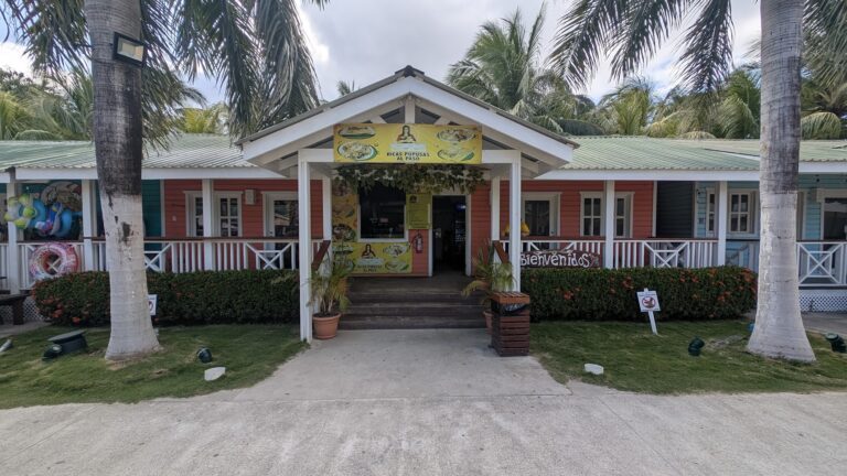 A colorful restaurant with a pink, blue, and green exterior surrounded by palm trees in Roatan, Honduras, featuring a wooden entrance with a "Bienvenidos" sign and a yellow sign advertising local traditional dishes, set in a tropical setting.