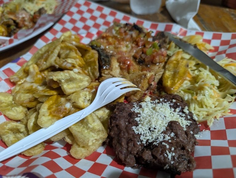 A tray of traditional American comfort foods featuring cheesy fries, a cheeseburger topped with pico de gallo, and a chocolate dessert with nuts, served on red and white checkered paper in Roatan, Honduras.