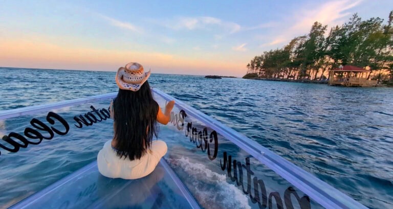 A woman wearing a cowboy hat sits on the transparent edge of a boat cruising in the waters near Roatan, Honduras, with lush trees and a pier visible along the shoreline at sunset.