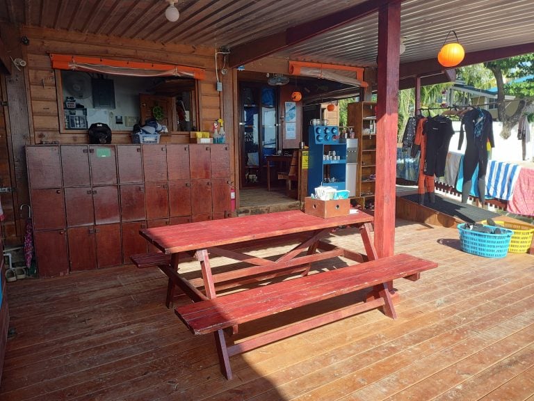 A wooden outdoor seating area with red picnic tables and hanging orange lanterns, featuring a clothing rack with wetsuits and tropical greenery in the background on Roatan, Honduras.