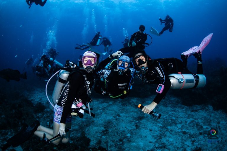 A group of scuba divers explores an underwater reef off Roatan in Honduras, with some divers posing for a photo while others swim nearby in clear blue water.