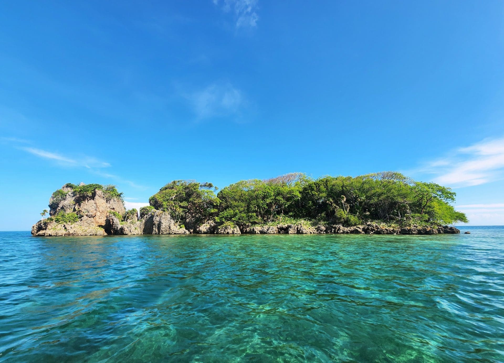 A lush green island with rocky cliffs covered in trees extends into clear turquoise waters under a bright blue sky in Roatan, Honduras.