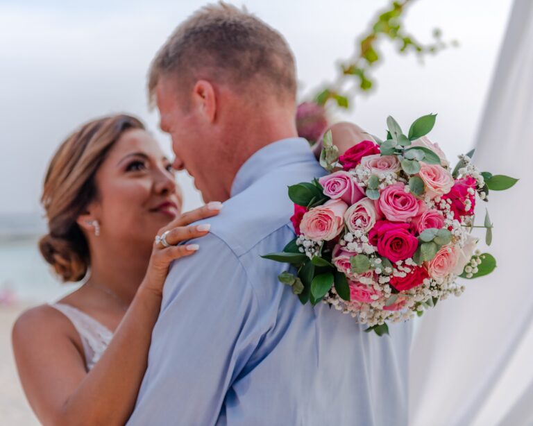 A bride and groom share a romantic moment on a beach in Roatan, Honduras, with the groom holding a large bouquet of pink and white roses, greenery, and baby's breath during their wedding ceremony.