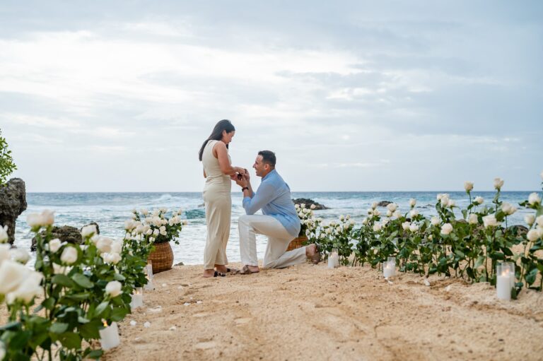 A couple exchanges vows during a beach wedding ceremony on Roatan, Honduras, with white roses and candles arranged in baskets and on the sand, ocean waves, and a cloudy sky in the background.