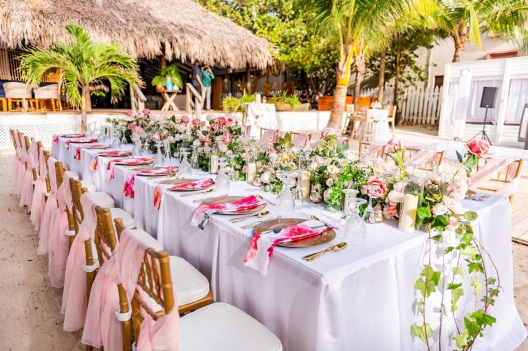 A decorated outdoor dining area with a long white table, pink chairs with tulle bows, floral centerpieces, and glassware, set under palm trees on a sandy surface in Roatan, Honduras.