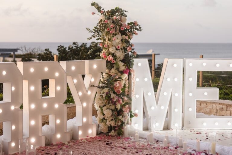 A wedding or event backdrop decorated with a large illuminated sign spelling "LOVE" and a tall floral arrangement of pink and white roses with green foliage, set against the ocean and sky in Roatan, Honduras.