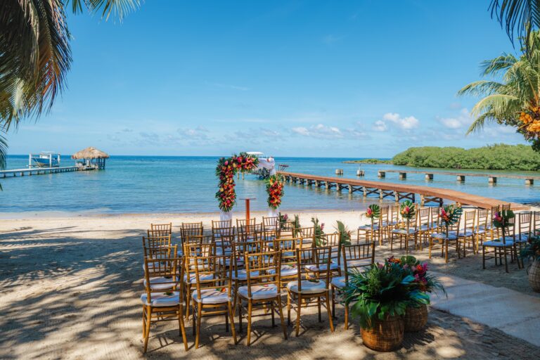 A beachfront wedding setup with chairs and floral arrangements on a sandy beach, overlooking the clear blue waters of Roatan, Honduras, with palm trees and a wooden pier extending into the ocean.