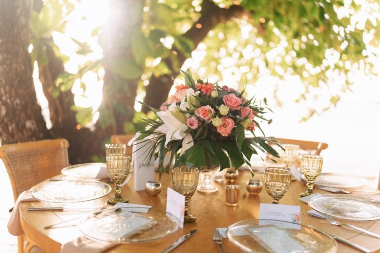 A round wooden dining table set with transparent glass plates, gold-rimmed glasses, silverware, and a large floral centerpiece featuring pink roses, white lilies, and greenery, situated outdoors under a leafy canopy in Roatan, Honduras.