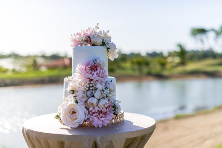 A wedding cake decorated with pink and white flowers and pearl accents is displayed on a table outdoors near a body of water on Roatan, Honduras.