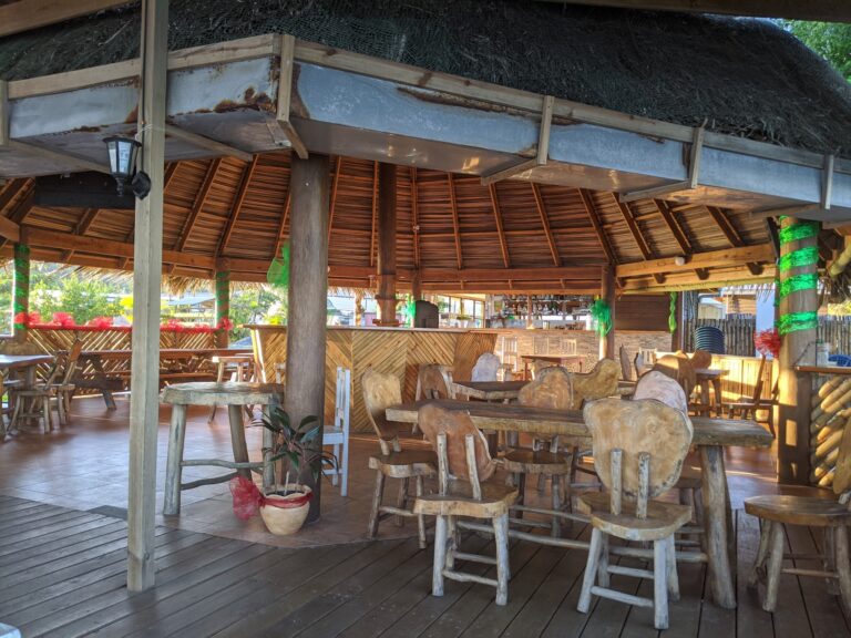 A thatched-roof outdoor bar area in Roatan, Honduras, features rustic wooden chairs and tables with colorful decorations and lush greenery surrounding the space.