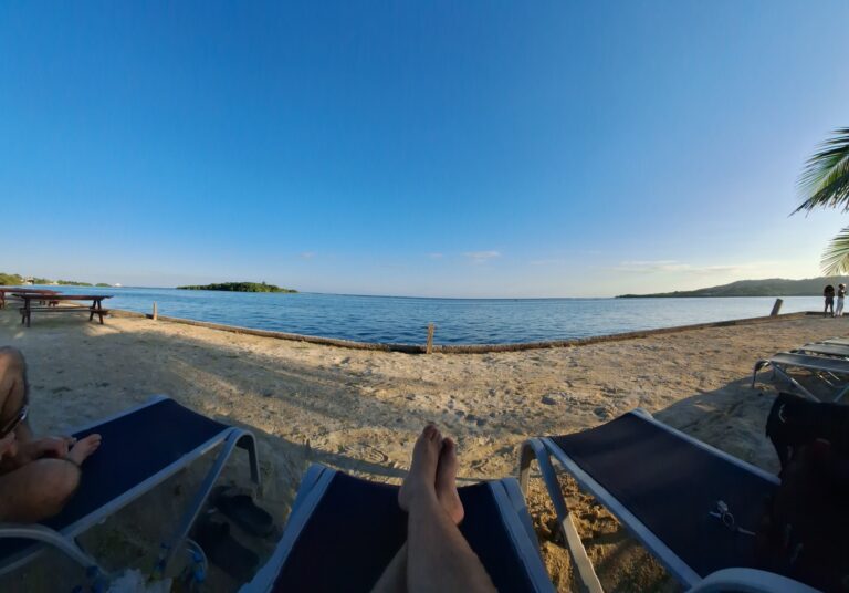 Relaxing view of a beach in Roatan, Honduras with lounge chairs, palm tree, calm water, and two people walking along the shoreline during sunset.