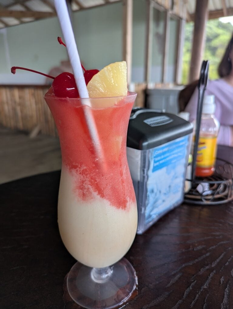 A layered tropical cocktail with red cherries and a pineapple wedge garnishes a tall glass on a dark wooden table at a restaurant in Roatan, Honduras, with a background of wooden and glass decor and a person sitting nearby.