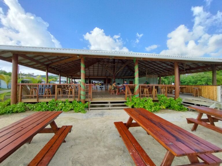A large open-air wooden pavilion with picnic tables and a view of green foliage, situated in Roatan, Honduras under a bright blue sky with scattered clouds.