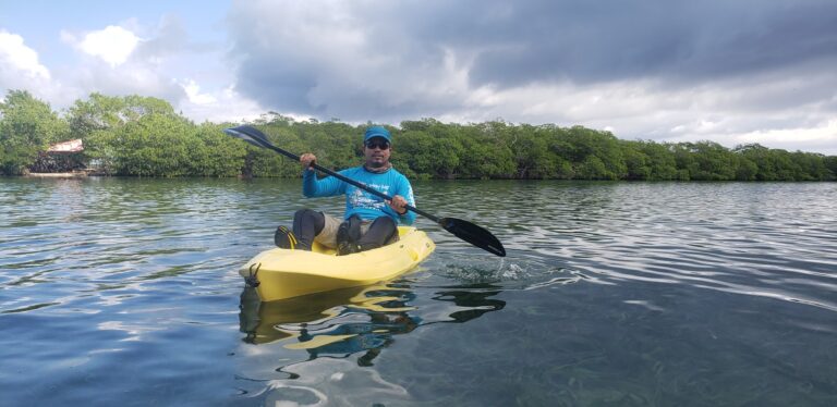 A man paddling a yellow kayak on calm waters near lush green mangroves on Roatan, Honduras, under a partly cloudy sky.