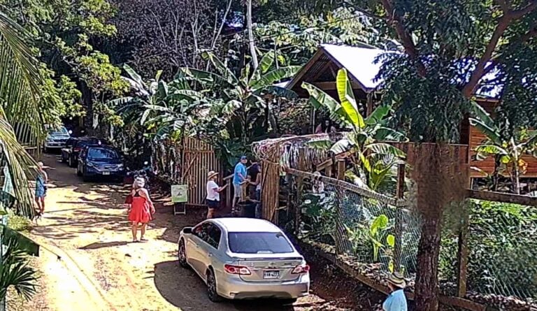 A group of people gathers outside a wooden building surrounded by lush tropical vegetation on Roatan, Bay Islands, Honduras, with several parked cars along a dirt road.