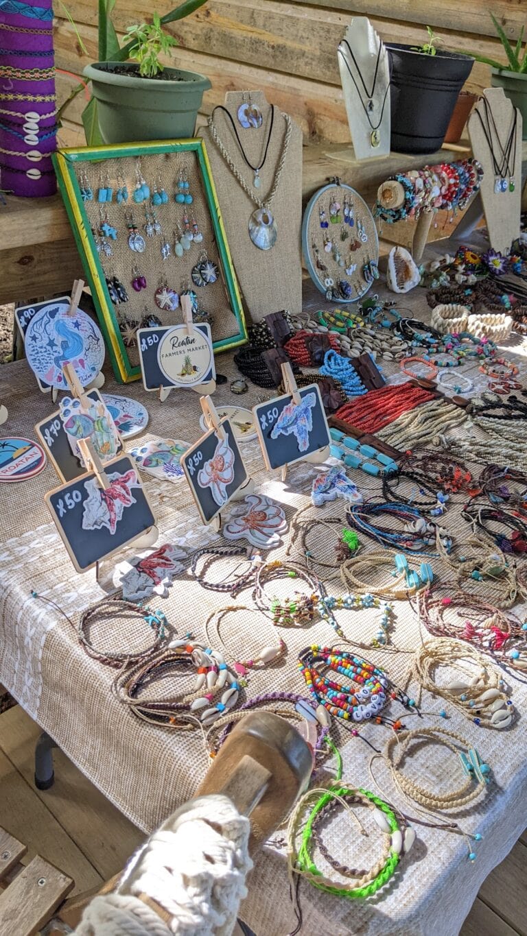 A display of handmade jewelry, including necklaces, bracelets, and earrings, arranged on tables and standees at a market stall in Roatan, Honduras, with potted plants and wooden walls in the background.