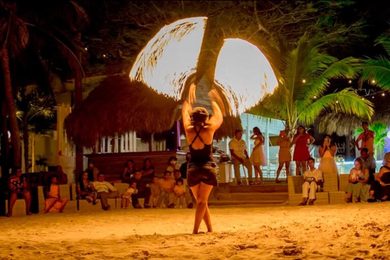A woman performs a fire dance on the sandy beach at night in Roatan, Honduras, with an audience seated and standing nearby, tropical palm trees, thatched-roof structures, and colorful lighting creating a lively island entertainment scene.