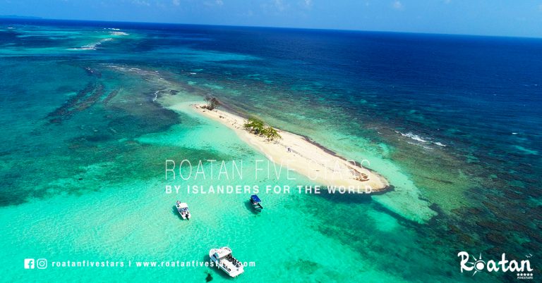Aerial view of a small, sandy island with a few trees surrounded by vibrant turquoise waters and coral reefs near Roatan, Honduras, with three boats anchored nearby.