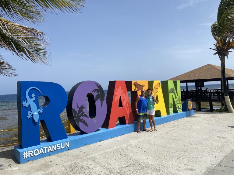 Colorful, large-lettered Roatan signage with a gecko graphic, located on the beach in Roatan, Honduras, with palm trees, a pavilion, and the ocean in the background.