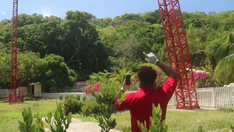 A man in a red shirt and white gloves takes a photo or video with his smartphone of a large red tower structure surrounded by lush green trees and colorful flowering plants in Roatan, Honduras.