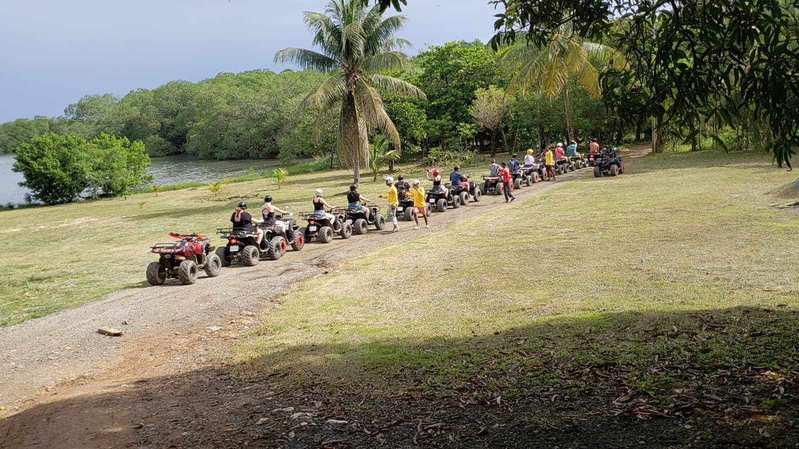 A group of people riding quad bikes along a dirt trail by a river with lush green trees and palm trees in Roatan, Honduras.