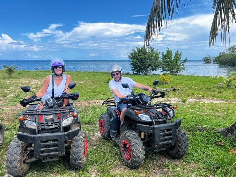 Two people riding all-terrain vehicles (ATVs) on a grassy coastal area with palm trees, a calm ocean, and a partly cloudy sky in the background on Roatan, Honduras.